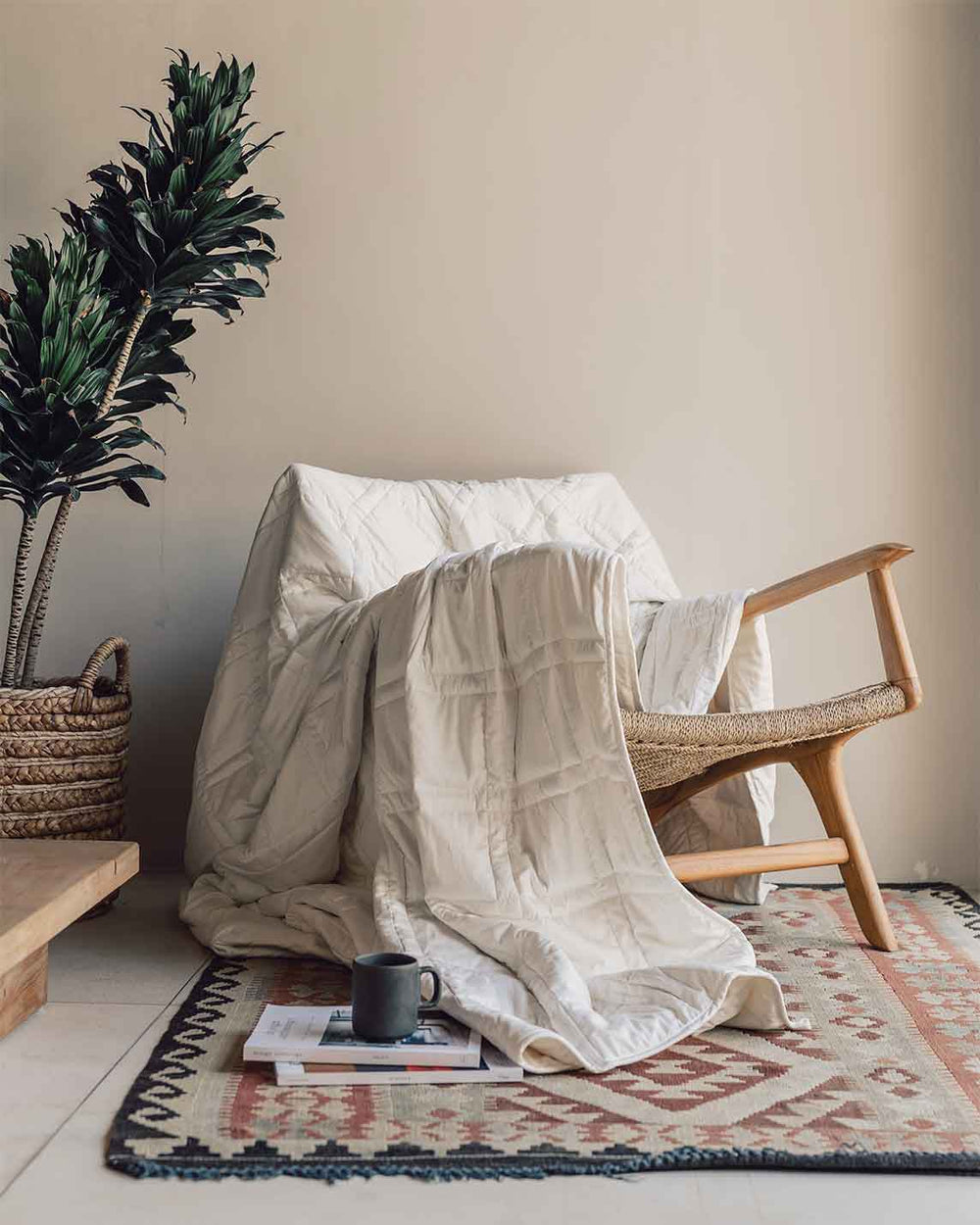 A cream weighted blanket draped over a woven-seat wood chair, its corner pooling on a patterned rug beside a mug and a stack of magazines.